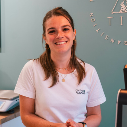 La gérante de l'épicerie Tipi à Theux, souriante derrière le comptoir, avec le logo du magasin en arrière-plan.