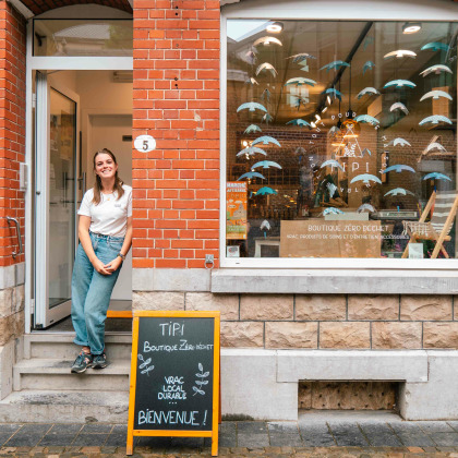 Façade de l'épicerie Tipi à Theux, avec la gérante debout devant l'entrée et une ardoise indiquant "Boutique zéro déchet" et "Bienvenue".