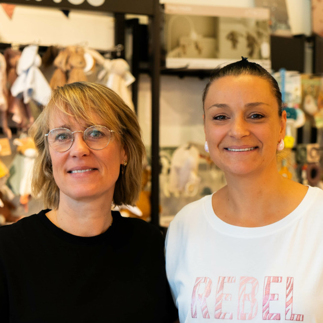 Les gérantes du magasin pour bébés Ptilou à Rocourt, souriantes devant des étagères remplies d'articles pour enfants.