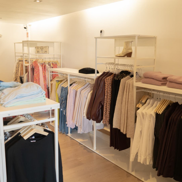 Portants de vêtements pour femmes et hommes dans le magasin Aries Clothing à Nandrin, avec des chemises, pulls et pantalons bien organisés.