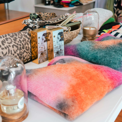 Articles de mode sur une table, incluant des sacs à motif léopard, des pulls colorés en laine, des flacons de parfum et d'autres accessoires exposés dans la boutique Intemporel à Ans