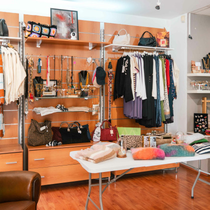 Intérieur de la boutique Intemporel, magasin de vêtements et accessoires avec des portants remplis de vêtements, des sacs à main, des bijoux, et des pulls colorés exposés sur une table au premier plan.