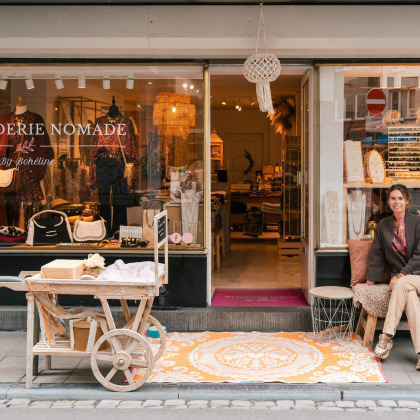 Devanture du magasin "Ma Penderie Nomade" à Liège avec Céline assise à l'entrée, entourée de vêtements bohème chic et d'une déco chaleureuse.