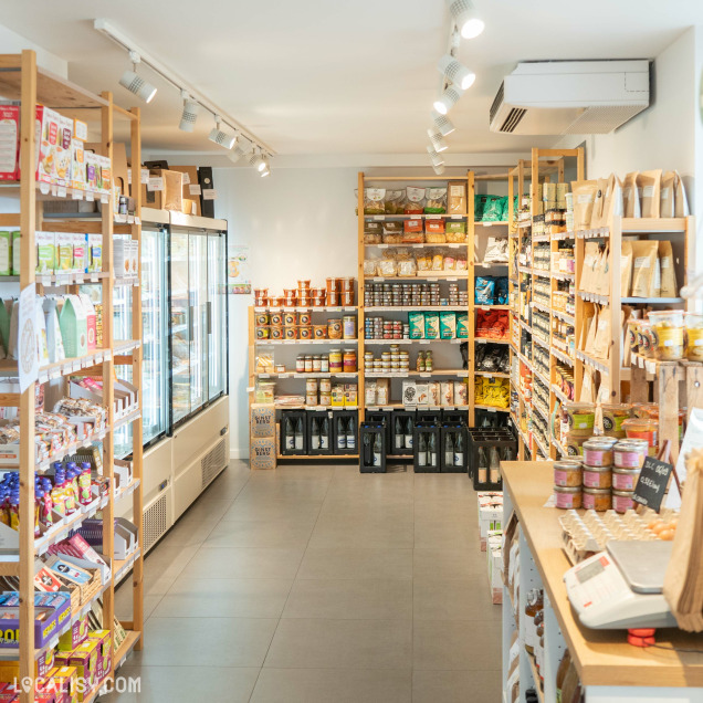 Intérieur du magasin Bio Au Comptoir Local à Beaufays, présentant des étagères remplies de produits alimentaires biologiques, des conserves, des snacks, des épices, ainsi qu'une section réfrigérée avec divers articles.