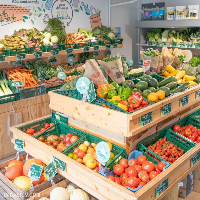 Section fruits et légumes du magasin Bio Au Comptoir Local à Beaufays, présentant des étagères remplies de légumes frais tels que des carottes, courgettes, poivrons, et tomates, avec une décoration murale sur la réduction des déchets.