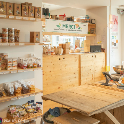 Comptoir du magasin Bio Au Comptoir Local à Beaufays, avec des étagères en bois remplies de produits locaux, un panneau de remerciement pour la réduction des déchets et des sacs en papier réutilisables.