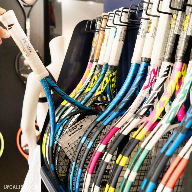 Collection de raquettes de tennis avec des poignées et des cordes de différentes couleurs, exposées sur un présentoir dans le magasin "Avantage Tennis" à Embourg