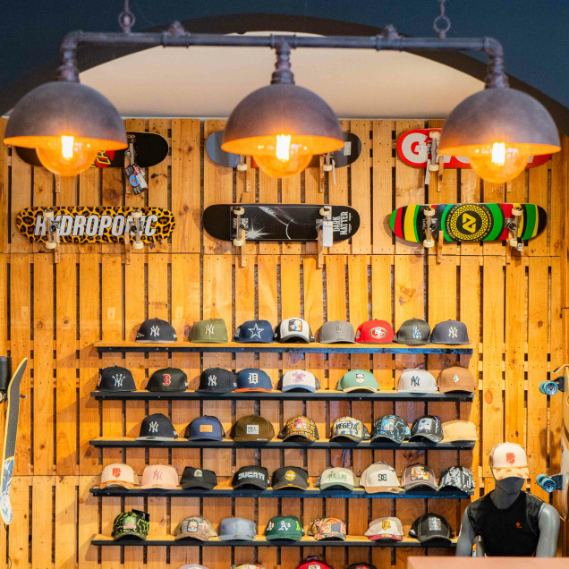 Intérieur du magasin “Bubba Ride” à Liège, vu à travers une entrée voûtée. Le mur du fond est en bois et présente plusieurs skateboards montés horizontalement. En dessous, des étagères exposent une collection de casquettes de différents designs.