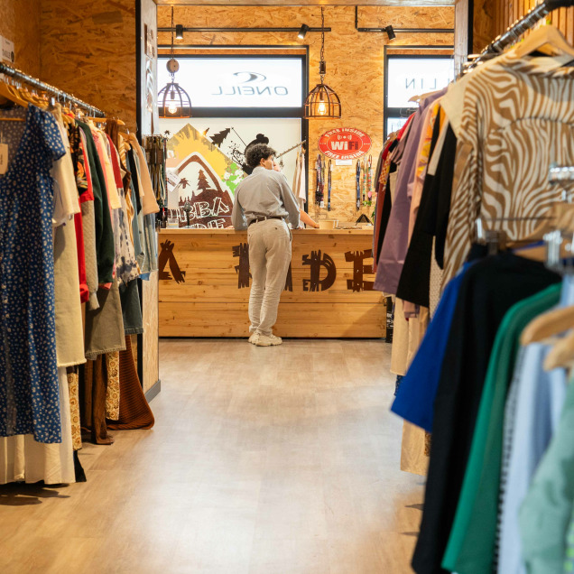 Magasin “Bubba Ride” à Liège avec sols et des murs en bois, divers vêtements exposés sur des portants et des cintres. Au centre, il y a un comptoir avec une personne derrière, de dos.