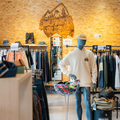 Chez “Bubba Ride” à Liège, on y voit divers vêtements exposés sur des portants et des étagères. Un panneau en bois avec le logo “BUBBA RIDE” est monté sur un mur en panneaux OSB, et il y a un mur en briques apparentes sur le côté.