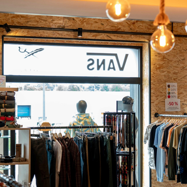 Intérieur du magasin “Bubba Ride” à Liège, avec des vêtements et accessoires exposés sur des portants et des étagères. Un design moderne avec des ampoules suspendues et un mur en bois en arrière-plan