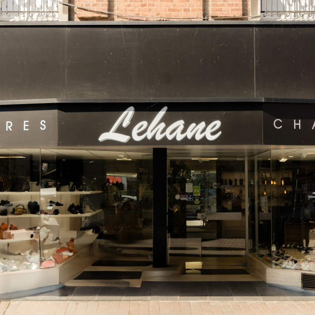 Devanture du magasin “Chaussures Lehane” à Visé. Le magasin a un auvent noir avec le nom “Lehane” écrit en lettres cursives blanches, encadré par le mot “Chaussures” de chaque côté. Les vitrines de chaque côté de l’entrée exposent une variété de chaussures pour différents âges et genres, disposées sur des étagères et des présentoirs.