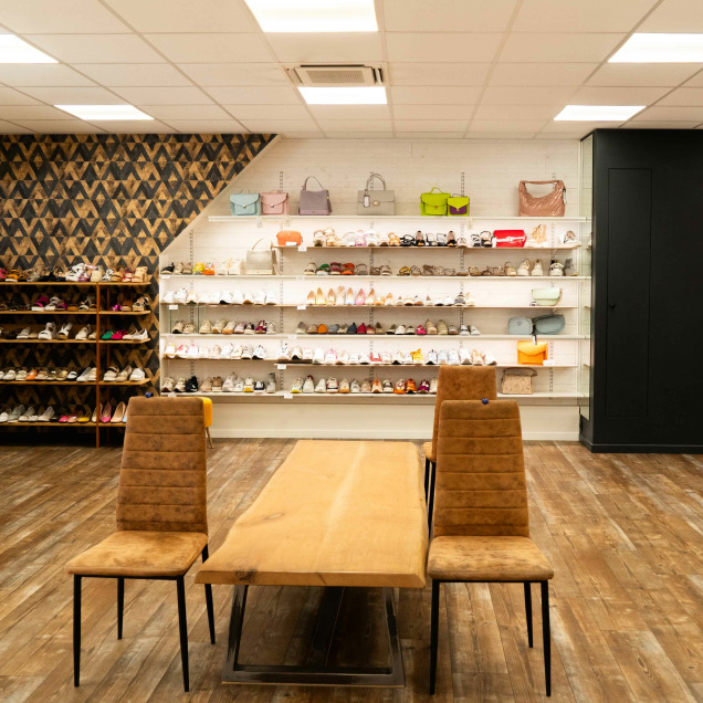 Intérieur du magasin “Chaussures Lehane” à Visé. Le magasin a un design lumineux et moderne avec des murs blancs et un sol en bois clair. À gauche, des étagères murales présentent diverses chaussures, tandis qu’à droite, des sacs à main sont disposés sur des étagères contre un fond à motifs.