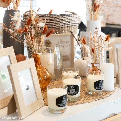 Divers articles de décoration exposés dans le magasin “Humeur du jour” ; des cadres, des bougies, un vase orange transparent avec des plantes séchées, et d’autres contenants décoratifs.