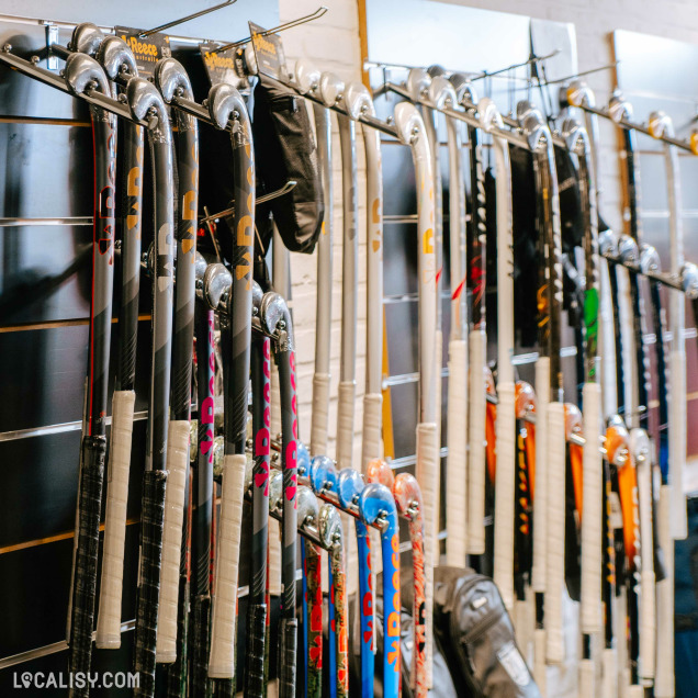 Variété de crosses de hockey alignées contre un mur, chacune avec des designs et des couleurs différents. La collection comprend des crosses avec des motifs noirs, blancs et divers, indiquant une large sélection disponible chez “Ikiba Sport” à Embourg.