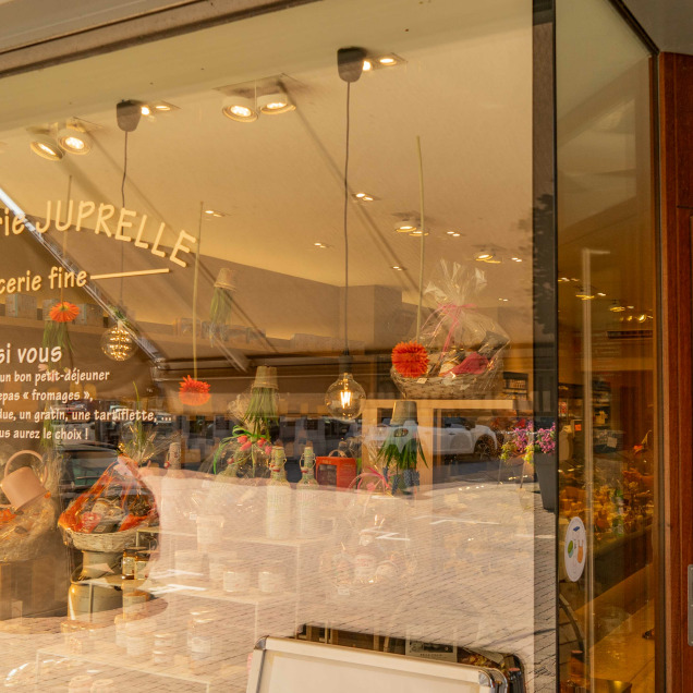 Devanture de la fromagerie “Maison Juprelle Cie” à Visé, une épicerie fine. La vitrine en verre reflète des bâtiments et le ciel de l’autre côté de la rue. L’intérieur semble bien éclairé avec des produits exposés, invitant les clients potentiels.