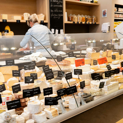 Intérieur de la fromagerie “Maison Juprelle Cie” à Visé avec une grande variété de fromages exposés dans une vitrine en verre. Deux personnes, probablement des employés, se tiennent derrière le comptoir.