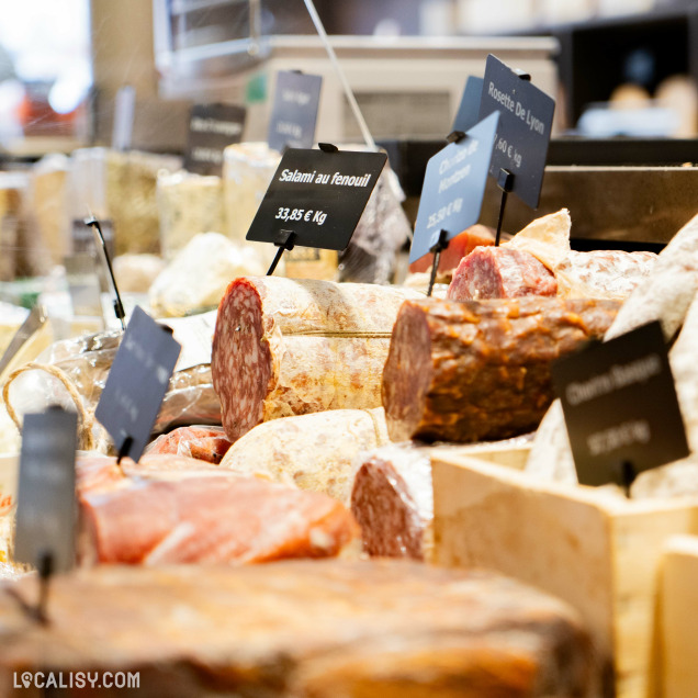 Sélection de fromages et de charcuteries exposés, avec des petites étiquettes noires indiquant les noms et les prix au kilogramme chez la “Maison Juprelle Cie” à Visé.