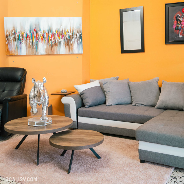 Salon chez Meubles Family à Hannut, avec un canapé d'angle gris, des tables basses en bois, et des œuvres d'art colorées accrochées sur un mur orange.