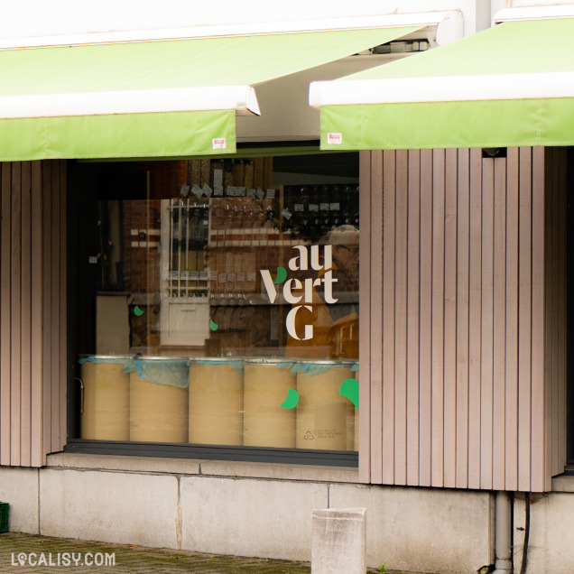 Devanture du magasin “Au Vert G” à Boncelles avec une vitrine encadrée par deux auvents verts et des panneaux en bois. Le nom du magasin est affiché en lettres minuscules sur la vitrine.