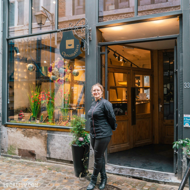 Devanture du magasin “Carré Noir” à Liège, avec une vitrine en verre exposant des produits et une façade décorée de fleurs. Une personne en vêtements sombres se tient devant, semblant entrer ou sortir du magasin.