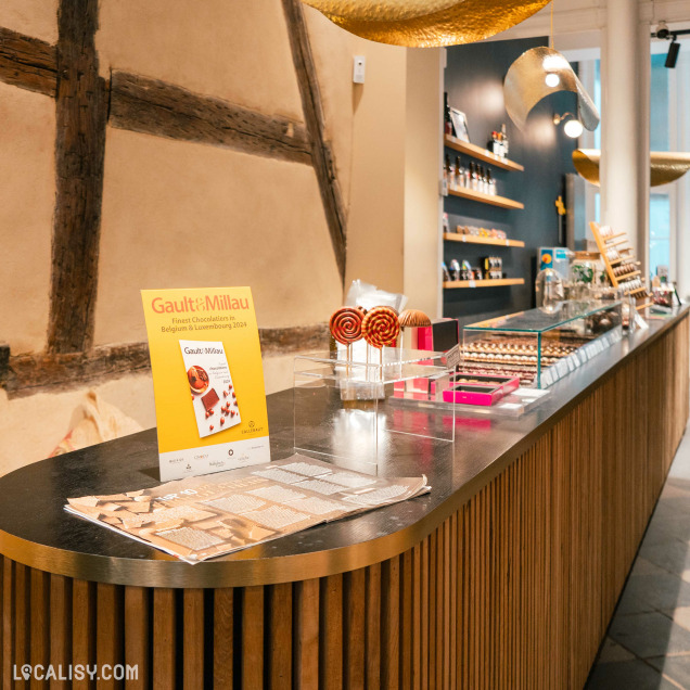 Comptoir présentant des pâtisseries et des douceurs, un guide “Gault&Millau” en évidence, et une décoration moderne avec des poutres en bois rustiques, dans le magasin "Carré Noir" à Liège