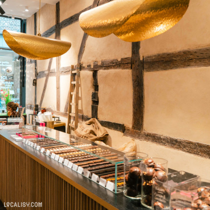 Intérieur du magasin “Carré Noir” à Liège, avec un comptoir de chocolats assortis, des cabosses dorées décoratives suspendues au-dessus, et un design mural rustique.