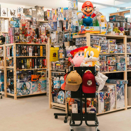 Une grande peluche de Mario est placée en évidence sur un chariot noir, qui contient également deux casquettes, l’une avec le logo “LA” et l’autre rouge sans logo visible au magasin "Computer Servces" à Theux