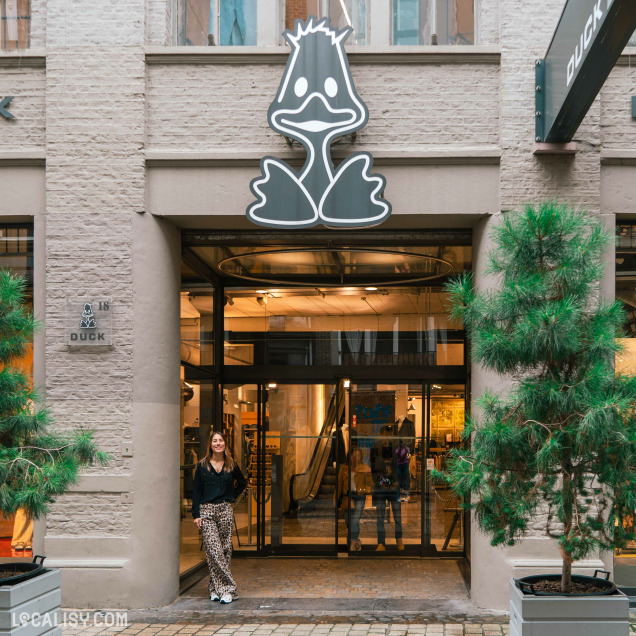 Devanture du magasin “Duck” à Liège présente un logo distinctif en forme de tête de canard au-dessus de l’entrée. Deux conifères en pot encadrent la façade, et une personne se tient à côté des portes vitrées.