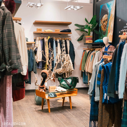 L’intérieur du magasin “Duck” à Liège présente une variété de vêtements sur cintres et étagères. Une table centrale en bois expose des accessoires, entourée de plantes et d’un tableau.