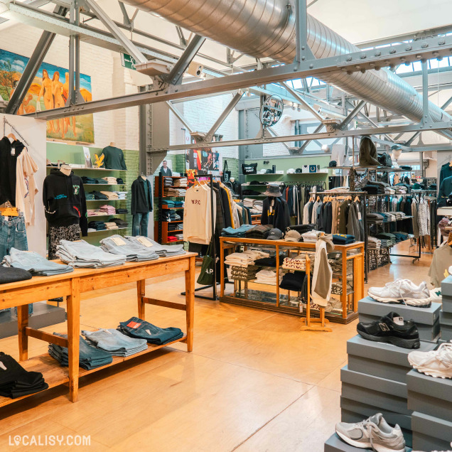 L’intérieur du magasin “Duck” à Liège présente des vêtements variés sur des tables et des étagères en bois, sous un éclairage lumineux.