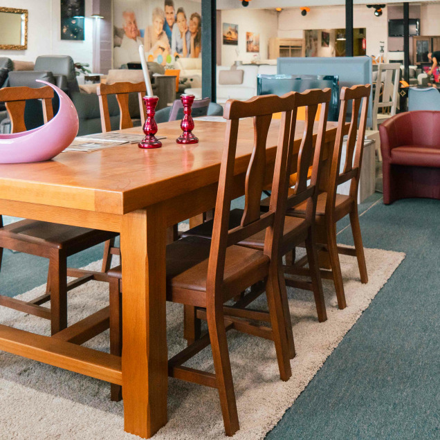 Une grande table en bois avec des chaises assorties est placée sur un tapis beige dans le showroom Quality Confort à Saint Georges-sur-Meuse, entourée de fauteuils en cuir et d'autres meubles d'exposition.