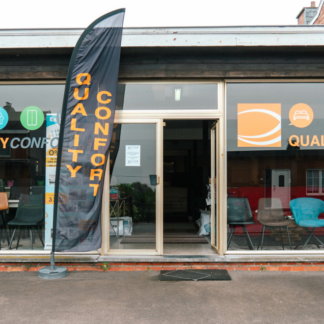 La façade du showroom Quality Confort à Saint Georges-sur-Meuse présente une large vitrine avec des chaises colorées exposées, ainsi qu'une enseigne visible avec le logo de l'enseigne et des icônes représentant différents meubles, accompagnée d'un drapeau publicitaire noir et orange à l'extérieur.