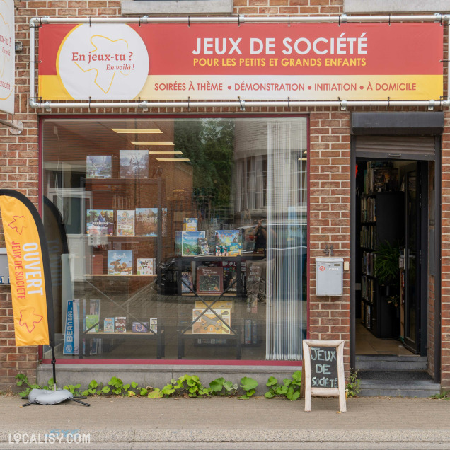 Devanture du magasin “En Jeux-Tu ? En Voilà !” à Saint-Georges-sur-Meuse. La vitrine affiche des jeux de société pour tous âges, avec des soirées à thème, démonstrations et initiations. Une pancarte indique que le magasin est ouvert.