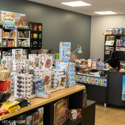 Intérieur du magasin “En Jeux-Tu ? En Voilà !” à Saint-Georges-sur-Meuse avec de nombreuses boîtes de jeux exposées sur des étagères et des tables. La variété des jeux suggère une offre adaptée à différents groupes d’âge et intérêts, allant des jeux familiaux aux jeux de stratégie plus complexes.