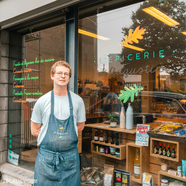 Devanture de "L’Épicerie d’Augustin" à Liège. La façade en verre affiche le nom du magasin en lettres blanches avec des motifs de feuilles vertes. Une personne portant un tablier se tient à l’entrée.