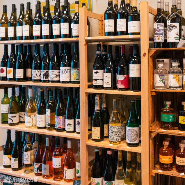 Étagères en bois remplies de bouteilles de vin et de spiritueux, avec des étiquettes variées, dans le magasin “L’Épicerie d’Augustin” à Liège.