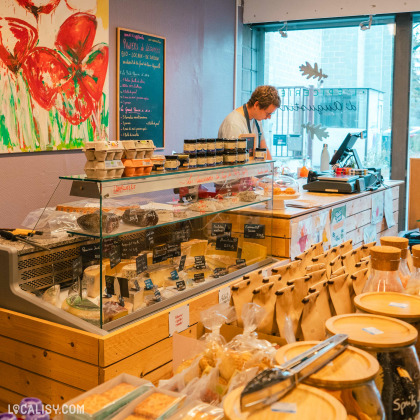 L’intérieur de “L’Épicerie d’Augustin” à Liège, on y voit divers produits alimentaires exposés sur des étagères et dans un comptoir en verre, avec des étiquettes manuscrites indiquant les noms et les prix des produits.