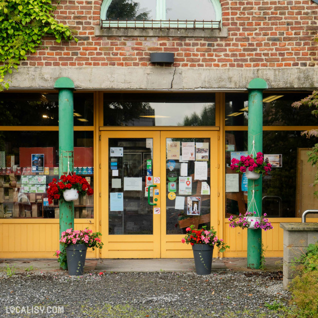 Devanture du magasin “La Prulhière” à Battice. L’entrée est encadrée par deux piliers verts et surmontée d’une façade en briques. Des plantes en pot avec des fleurs colorées ajoutent une touche accueillante à l’entrée.