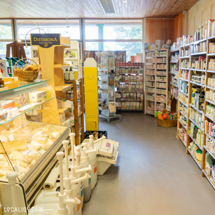 Des étagères en bois avec divers produits, un présentoir réfrigéré avec des fromages, et des paniers remplis de marchandises à l’intérieur du magasin “La Prulhière” à Battice.