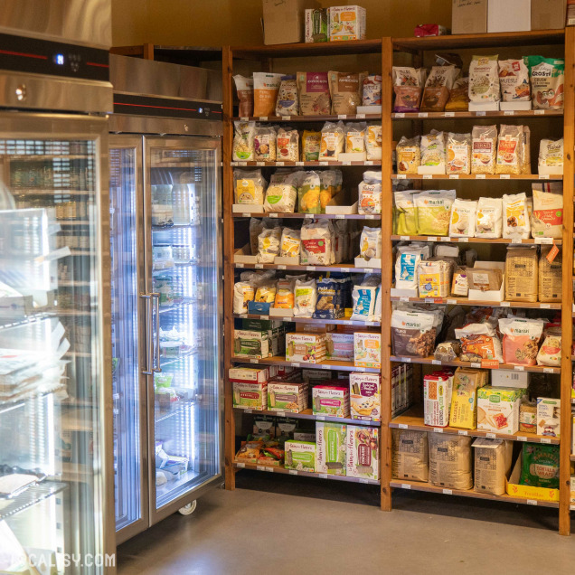 L’intérieur du magasin “La Prulhière” à Battice avec des étagères en bois remplies de divers produits emballés et un présentoir réfrigéré sur la gauche.