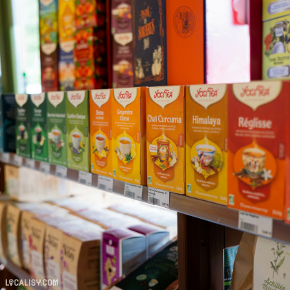 Une étagère dans le magasin “La Prulhière” à Battice, remplie de boîtes de thé colorées de la marque “Yogi Tea” avec des saveurs comme ‘Classic’, ‘Ginger Lemon’, ‘Chai Curcuma’, ‘Himalaya’, et ‘Réglisse’.