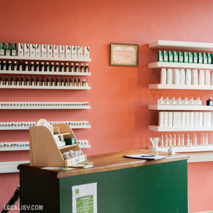 L’intérieur du magasin “Martine Van” à Esneux présente un comptoir en bois avec une façade verte, surmonté de quelques papiers et d’un petit panneau blanc. Derrière le comptoir, contre un mur orange, se trouvent des étagères blanches bien organisées avec divers flacons et contenants.