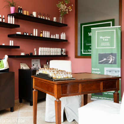 Une table en bois au centre avec plusieurs petits flacons, des étagères murales exposant divers produits, et une affiche verte avec le texte “Martine Van” au magasin “Martine Van” à Esneux.