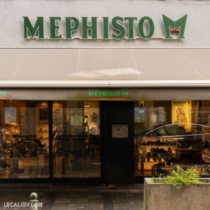 Devanture du magasin “Mephisto Shop” à Liège. Le nom du magasin est affiché en grandes lettres blanches sur un fond vert avec un logo rouge en forme de “M” au-dessus. La porte d’entrée noire porte le numéro 15, et deux plantes en pot sont visibles de chaque côté de l’entrée.