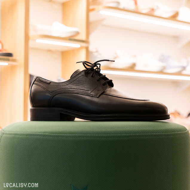 Une chaussure noire à lacets exposée sur un piédestal vert avec un emblème en forme d’étoile. En arrière-plan, des étagères présentent d’autres chaussures au magasin “Mephisto Shop” à Liège