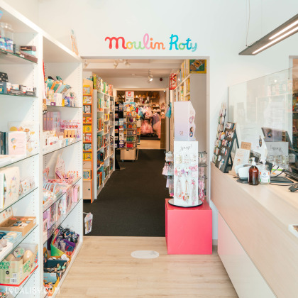 Variété de produits tels que des jouets, des livres et des vêtements. Une enseigne visible au fond porte le texte “Moulin Roty” au magasin “Les Z’arsouilles” à Theux, suggérant une section dédiée à cette marque connue pour ses jouets en peluche et sa décoration pour enfants.