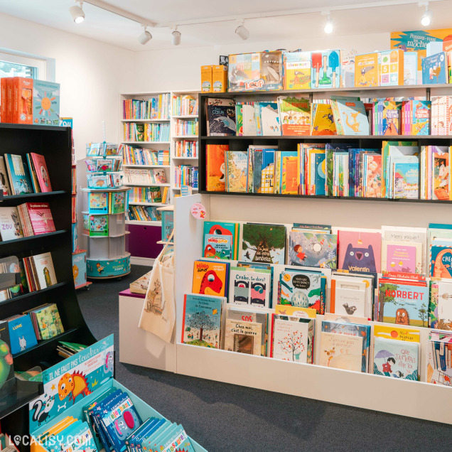 L’intérieur du magasin “Les Z’arsouilles” à Theux rempli de livres pour enfants, avec des étagères colorées et des couvertures de livres attrayantes.