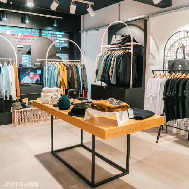 L’intérieur du magasin “MILK” à Liège a un design moderne avec des portants et des étagères bien organisés, exposant divers vêtements comme des chemises, des vestes et des chapeaux.