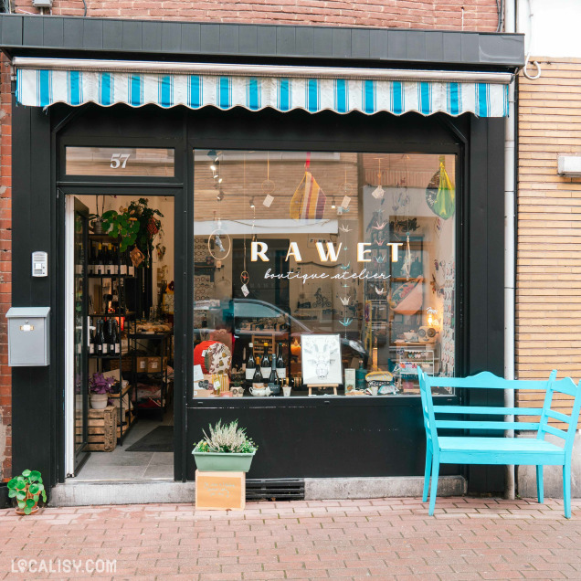 Devanture du magasin “Rawet” à Angleur. La façade est noire avec un auvent bleu et blanc rayé. La vitrine expose divers articles, et une plante en pot ainsi qu’un banc bleu sont placés devant le magasin.